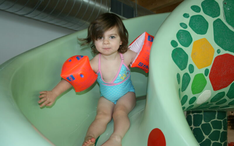Ein kleines Kind, das orangefarbene Schwimmflügel und einen Badeanzug trägt, rutscht eine grüne Indoor-Wasserrutsche mit einem Schildkrötenmosaik hinunter.