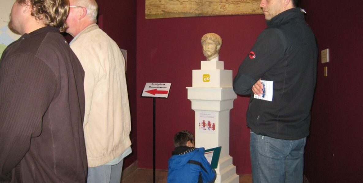 Vier Personen in einem Museumsraum mit roten Wänden; drei Erwachsene stehen neben einer Statuenbüste, während ein Kind in einer blauen Jacke sitzt und auf einem Klemmbrett schreibt oder zeichnet.