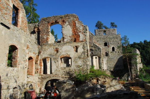 Ruinen eines alten Steingebäudes mit großen Fensteröffnungen, freiliegendem Mauerwerk und umliegendem Grün unter strahlend blauem Himmel.