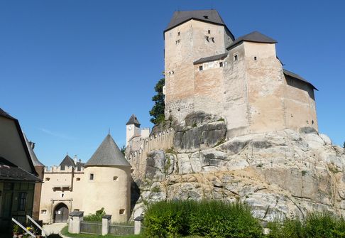 Eine steinerne Burg mit mehreren Türmen und Mauern thront auf einem felsigen Hügel unter strahlend blauem Himmel, umgeben von Grün.