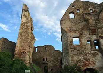 Ruinen einer alten Steinburg mit teilweise eingestürzten Mauern und Türmen, umgeben von Grün, unter einem teilweise bewölkten Himmel.