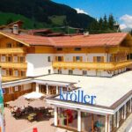 Ein dreistöckiges Alpenhotel mit Holzbalkonen und rotem Dach, mit einer Außenterrasse und einem Schild mit der Aufschrift "Kröller", in einer Berglandschaft gelegen.