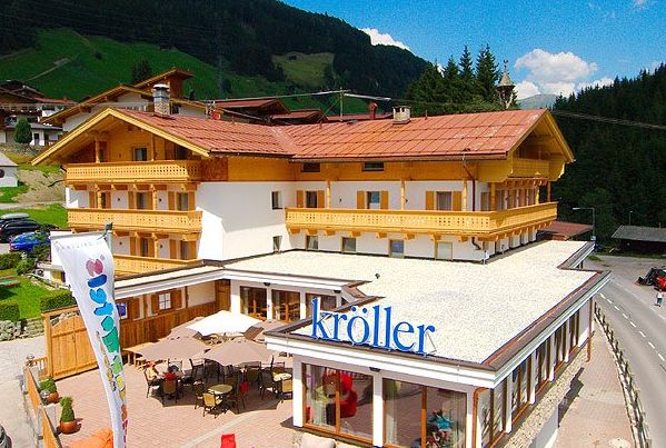 Ein dreistöckiges Alpenhotel mit Holzbalkonen und rotem Dach, mit einer Außenterrasse und einem Schild mit der Aufschrift "Kröller", in einer Berglandschaft gelegen.
