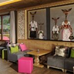 Gemütliches Wohnzimmer mit moderner Einrichtung, grauen Sofas, bunten Kissen und Hockern, Holzakzenten und großen Gemälden von anthropomorphen Hirschen in traditioneller Kleidung an der Wand.