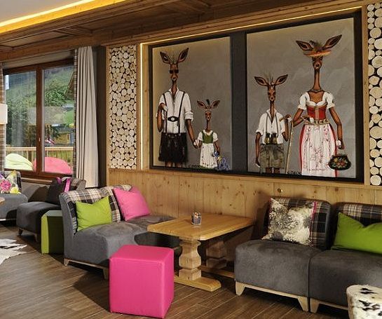 Gemütliches Wohnzimmer mit moderner Einrichtung, grauen Sofas, bunten Kissen und Hockern, Holzakzenten und großen Gemälden von anthropomorphen Hirschen in traditioneller Kleidung an der Wand.