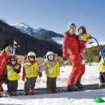 Zwei Erwachsene und fünf Kinder in Skiausrüstung stehen im Schnee; die Kinder tragen gelbe Westen und Helme, im Hintergrund sind Berge und Bäume zu sehen.