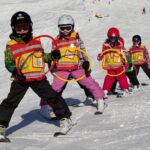 Fünf Kinder mit Helmen und Sicherheitswesten fahren in einer Reihe auf einer verschneiten Piste Ski, jedes hält einen farbigen Ring.