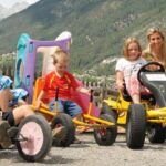 Vier Kinder und ein Erwachsener sitzen auf Gokarts und Spielzeug im Freien mit Bergen und Grün im Hintergrund.