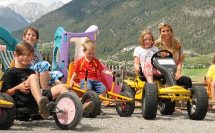 Vier Kinder und ein Erwachsener sitzen auf Gokarts und Spielzeug im Freien mit Bergen und Grün im Hintergrund.