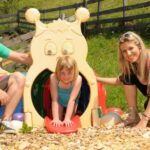 Ein Mann und eine Frau hocken neben einem lächelnden Mädchen, das in einem raupenförmigen Spielplatztunnel im Freien spielt.