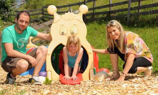 Ein Mann und eine Frau hocken neben einem lächelnden Mädchen, das in einem raupenförmigen Spielplatztunnel im Freien spielt.