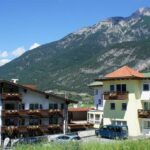 Eine Gruppe von Gebäuden im alpinen Stil mit Blumenkästen liegt am Fuße eines grünen Berges unter einem klaren blauen Himmel.
