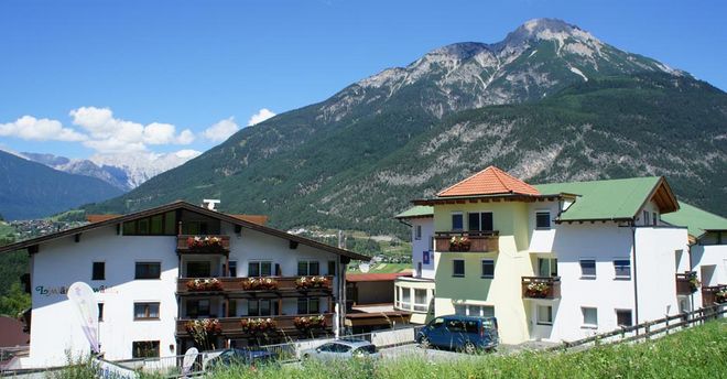 Eine Gruppe von Gebäuden im alpinen Stil mit Blumenkästen liegt am Fuße eines grünen Berges unter einem klaren blauen Himmel.