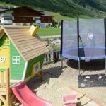 Ein kleines grünes Spielhaus mit einer Rutsche und einem Sandkasten steht neben einem runden Trampolin mit einem Sicherheitsnetz auf einem Spielplatz im Freien mit grasbewachsenen Hügeln im Hintergrund.