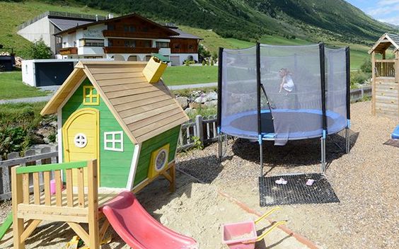 Ein kleines grünes Spielhaus mit einer Rutsche und einem Sandkasten steht neben einem runden Trampolin mit einem Sicherheitsnetz auf einem Spielplatz im Freien mit grasbewachsenen Hügeln im Hintergrund.