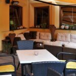 Sitzbereich im Freien mit einem weißen Tisch, schwarzen Korbstühlen mit gelben Kissen und einem gepolsterten Ecksofa vor einer gelben Wand mit Fenstern und Topfpflanzen.