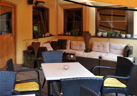 Sitzbereich im Freien mit einem weißen Tisch, schwarzen Korbstühlen mit gelben Kissen und einem gepolsterten Ecksofa vor einer gelben Wand mit Fenstern und Topfpflanzen.