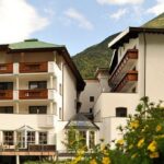 Ein mehrstöckiges Alpenhotel mit Holzbalkonen, weißen Wänden und grünem Dach, umgeben von Blumen und Bergen unter einem teilweise bewölkten Himmel.