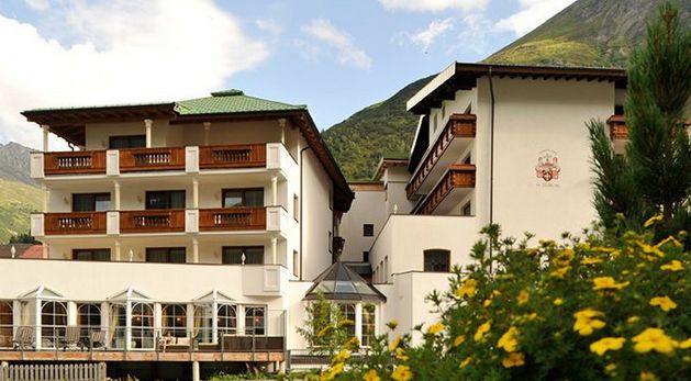 Ein mehrstöckiges Alpenhotel mit Holzbalkonen, weißen Wänden und grünem Dach, umgeben von Blumen und Bergen unter einem teilweise bewölkten Himmel.