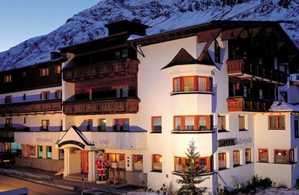 Ein mehrstöckiges alpines Hotel mit Holzbalkonen und weißen Wänden vor dem Hintergrund schneebedeckter Berge wird in der Abenddämmerung mit einer Weihnachtsmannfigur am Eingang beleuchtet.