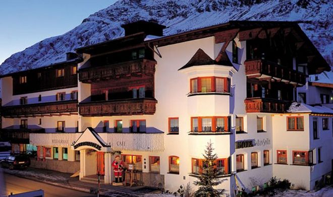 Ein mehrstöckiges alpines Hotel mit Holzbalkonen und weißen Wänden vor dem Hintergrund schneebedeckter Berge wird in der Abenddämmerung mit einer Weihnachtsmannfigur am Eingang beleuchtet.