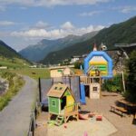 Ein kleiner Spielplatz mit Spielhäusern und einer Hüpfburg liegt neben einem Bach und einer Berglandschaft unter einem teilweise bewölkten Himmel.