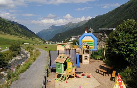 Ein kleiner Spielplatz mit Spielhäusern und einer Hüpfburg liegt neben einem Bach und einer Berglandschaft unter einem teilweise bewölkten Himmel.