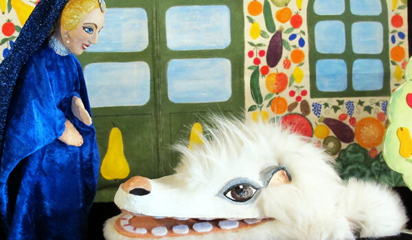 Eine Puppe mit blauem Mantel steht einer weißen, pelzigen Kreatur mit großen Augen und Zähnen gegenüber, die vor einem bunten, gemalten Hintergrund mit Fenstern und Obstbäumen steht.