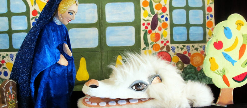 Eine Puppe mit blauem Mantel steht einer weißen, pelzigen Kreatur mit großen Augen und Zähnen gegenüber, die vor einem bunten, gemalten Hintergrund mit Fenstern und Obstbäumen steht.