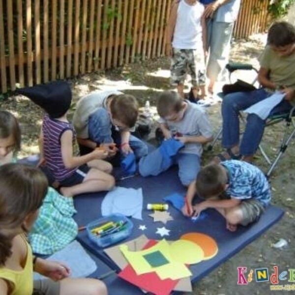 Mehrere Kinder sitzen und knien im Freien auf einer Matte und basteln mit Papier, Buntstiften und Klebstoff. Ein Erwachsener sitzt in der Nähe und schaut zu.