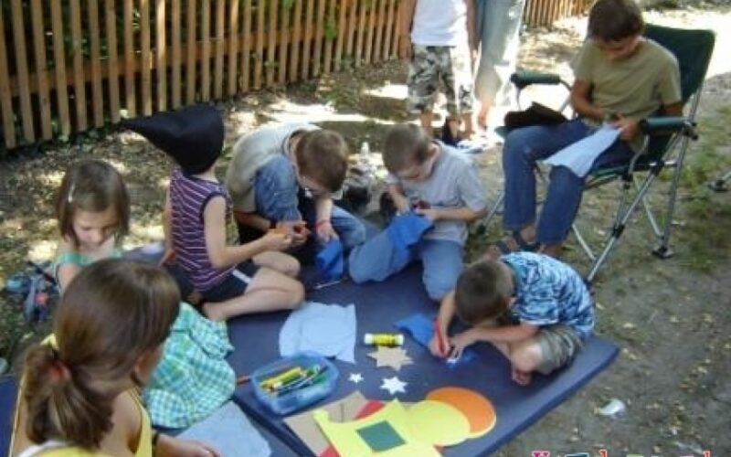 Mehrere Kinder sitzen und knien im Freien auf einer Matte und basteln mit Papier, Buntstiften und Klebstoff. Ein Erwachsener sitzt in der Nähe und schaut zu.