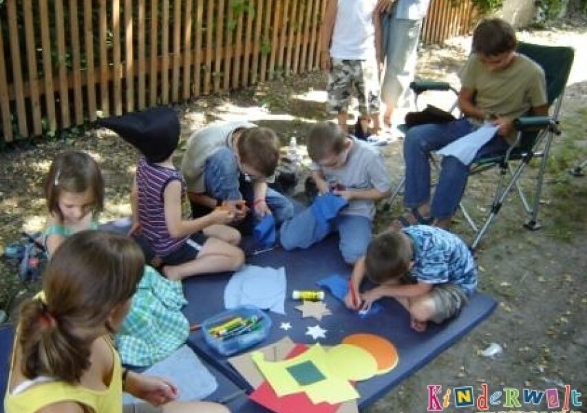 Mehrere Kinder sitzen und knien im Freien auf einer Matte und basteln mit Papier, Buntstiften und Klebstoff. Ein Erwachsener sitzt in der Nähe und schaut zu.