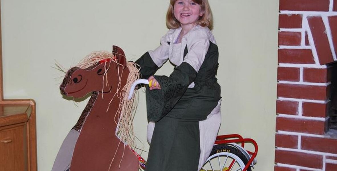 Ein junges Mädchen in einem Kleid sitzt auf einem Fahrrad, das mit einer Pappfigur eines Pferdes verziert ist, und lächelt in der Nähe eines gemauerten Kamins.