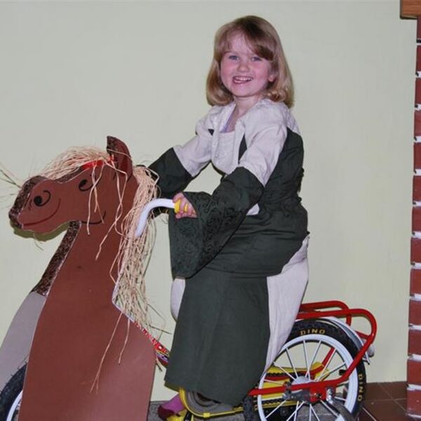Ein junges Mädchen in einem Kleid sitzt auf einem Fahrrad, das mit einer Pappfigur eines Pferdes verziert ist, und lächelt in der Nähe eines gemauerten Kamins.