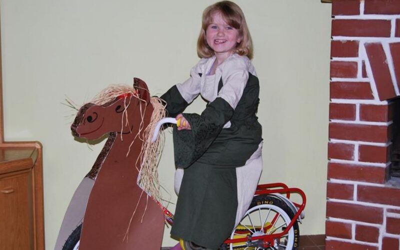Ein junges Mädchen in einem Kleid sitzt auf einem Fahrrad, das mit einer Pappfigur eines Pferdes verziert ist, und lächelt in der Nähe eines gemauerten Kamins.