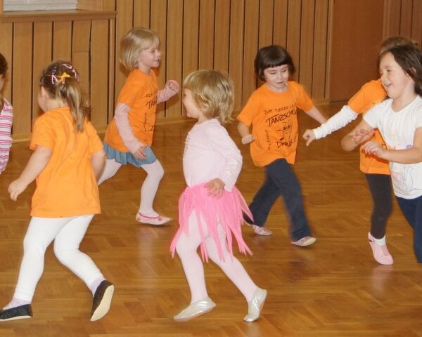 Eine Gruppe kleiner Kinder spielt und rennt zusammen in einer Turnhalle, einige tragen orangefarbene Hemden und eines ein rosa Tutu.