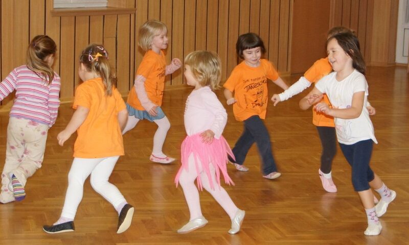 Eine Gruppe kleiner Kinder spielt und rennt zusammen in einer Turnhalle, einige tragen orangefarbene Hemden und eines ein rosa Tutu.