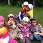 Sechs Kinder sitzen und knien im Gras, lächeln und streicheln einen braun-weißen Hund, im Hintergrund sind Bäume und Grünpflanzen zu sehen.