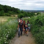 Eine Gruppe von Menschen, darunter Kinder und ein Hund, geht an einem bedeckten Tag einen unbefestigten Weg durch grüne Vegetation entlang.