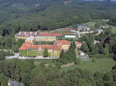 Luftaufnahme eines großen Gebäudekomplexes mit roten Dächern, umgeben von Bäumen und bewaldeten Flächen.