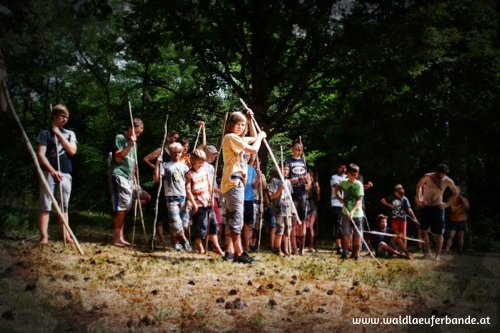 Eine Gruppe von Kindern und Jugendlichen steht im Freien auf einer Lichtung, jeder hält einen langen Holzstab in der Hand, umgeben von Bäumen im Tageslicht.