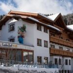 Ein mehrstöckiges Alpenhotel mit Holzbalkonen, schneebedecktem Dach und Cartoon-Maskottchen-Dekoration an der Fassade, umgeben von verschneiten Bäumen und Bergen.