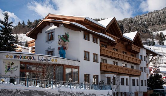 Ein mehrstöckiges Alpenhotel mit Holzbalkonen, schneebedecktem Dach und Cartoon-Maskottchen-Dekoration an der Fassade, umgeben von verschneiten Bäumen und Bergen.