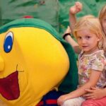 Zwei kleine Kinder sitzen auf einem großen, bunten Raupenmaskottchen in einem Spielbereich und lächeln in die Kamera.