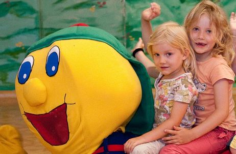 Zwei kleine Kinder sitzen auf einem großen, bunten Raupenmaskottchen in einem Spielbereich und lächeln in die Kamera.