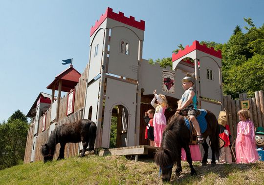 Verkleidete Kinder spielen in der Nähe eines Spielplatzes, auf dem eine Burg aufgebaut ist; ein Kind reitet auf einem Pony, und ein anderes Pony grast auf der Wiese neben ihnen.