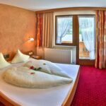 Ein Hotelzimmer mit einem Doppelbett, einem rot gestreiften Sofa, einem Holzstuhl, zwei Fenstern mit Vorhängen und einem roten Teppichboden.