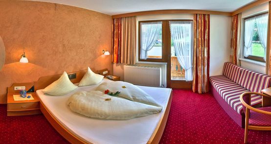 Ein Hotelzimmer mit einem Doppelbett, einem rot gestreiften Sofa, einem Holzstuhl, zwei Fenstern mit Vorhängen und einem roten Teppichboden.