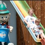 Zwei Kinder rutschen eine bunte Spielplatzrutsche hinunter, die neben einer großen, karikaturistischen Statue eines Jungen mit grünem Hut und blauem Eimer steht.