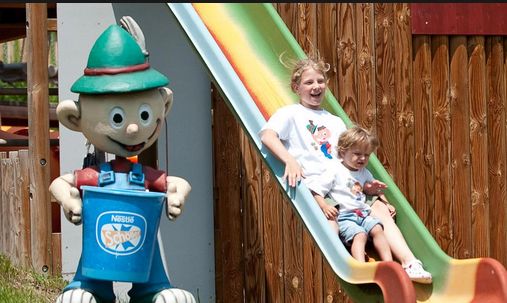 Zwei Kinder rutschen eine bunte Spielplatzrutsche hinunter, die neben einer großen, karikaturistischen Statue eines Jungen mit grünem Hut und blauem Eimer steht.